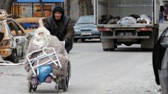 Un hombre empuja una silla de ruedas con una anciana y, al fondo, un camin con cadveres en Maripol