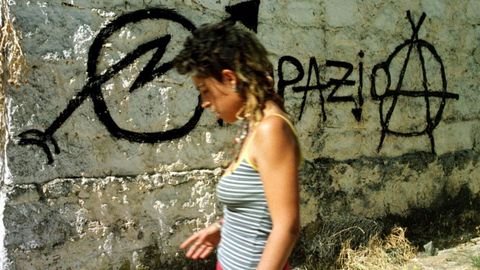 Una mujer ocupa pasa frente a un muro con el smbolo del movimiento y otro anarquista.