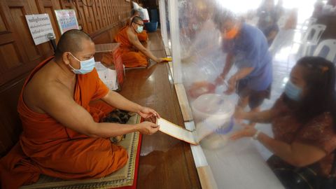 Monjes budistas, protegidos con mascarillas y una cortina de plstico reciben ofrendas de sus devotos en un monasterio de Bangkok, Tailandia