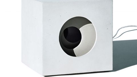Lámpara de Hormigón de Es Devlin. Una lámpara en forma de cubo, moldeada en hormigón sólido, que presenta el gesto de iris en capas con el que Devlin trabaja habitualmente en sus instalaciones cinéticas a gran escala. Una de las caras de la lámpara está grabada con una abertura vertical de luz, otro de los motivos recurrentes de la artista; otra cara está tallada con una serie de vacíos circulares concéntricos. Reducción en escala de las monumentales esculturas giratorias de Devlin, la lámpara traslada elementos de la instalación pública a un formato doméstico. Producida en una edición limitada de 20 unidades.