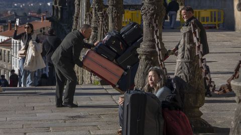 Un trabajador se afana en trasladar equipajes hacia el Hostal de los Reyes Catlicos mientras una turista descansa sentada en el Obradoiro