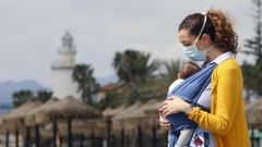 Una mujer protegida con una mascarilla da un paseo con su beb� en la playa de La Malagueta