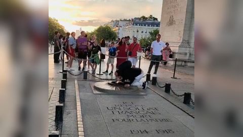 El hombre, de rodillas, encendindose el cigarrillo con la llama del monumento.