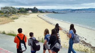 Un grupo de jóvenes caminan por el paseo de Cangas mientras observan la playa de Rodeira, completamente desierta bajo un cielo de nubes bajas.